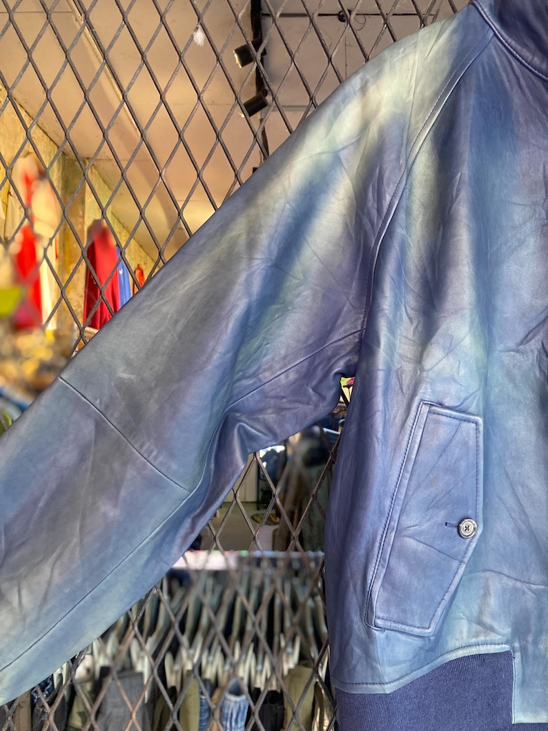 May include: A blue leather jacket with a distinctive tie-dye pattern. The jacket has a buttoned pocket and a ribbed hem. The sleeves display a gradient of blue and white tones. A fashionable outerwear garment.
