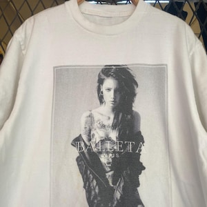 May include: White crew neck t-shirt featuring a black and white graphic of a person with long hair and tattoos, wearing a jacket. The word "BALLETA" is printed on the shirt.