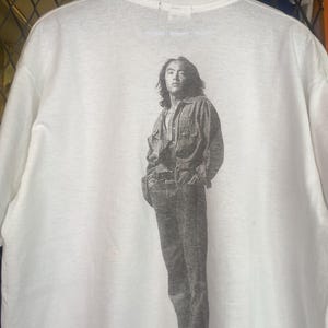Pode incluir: Camiseta branca com uma imagem a preto e branco de uma pessoa com camisa de ganga e calças de ganga. A imagem está centrada nas costas da camiseta. A camiseta tem gola redonda e mangas curtas.