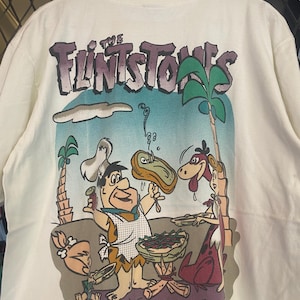 May include: White t-shirt featuring a cartoon image of the Flintstones family enjoying a barbecue. The shirt has the words "The Flintstones" printed in purple on the back.