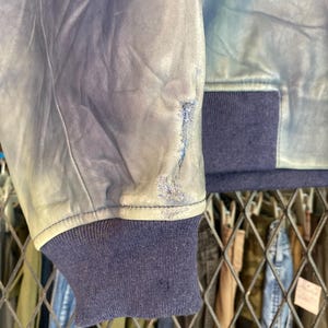 May include: Close-up of a distressed blue leather jacket sleeve. The jacket has a navy blue ribbed cuff and a light blue leather exterior with visible wear, giving a vintage look. The sleeve shows signs of wear.