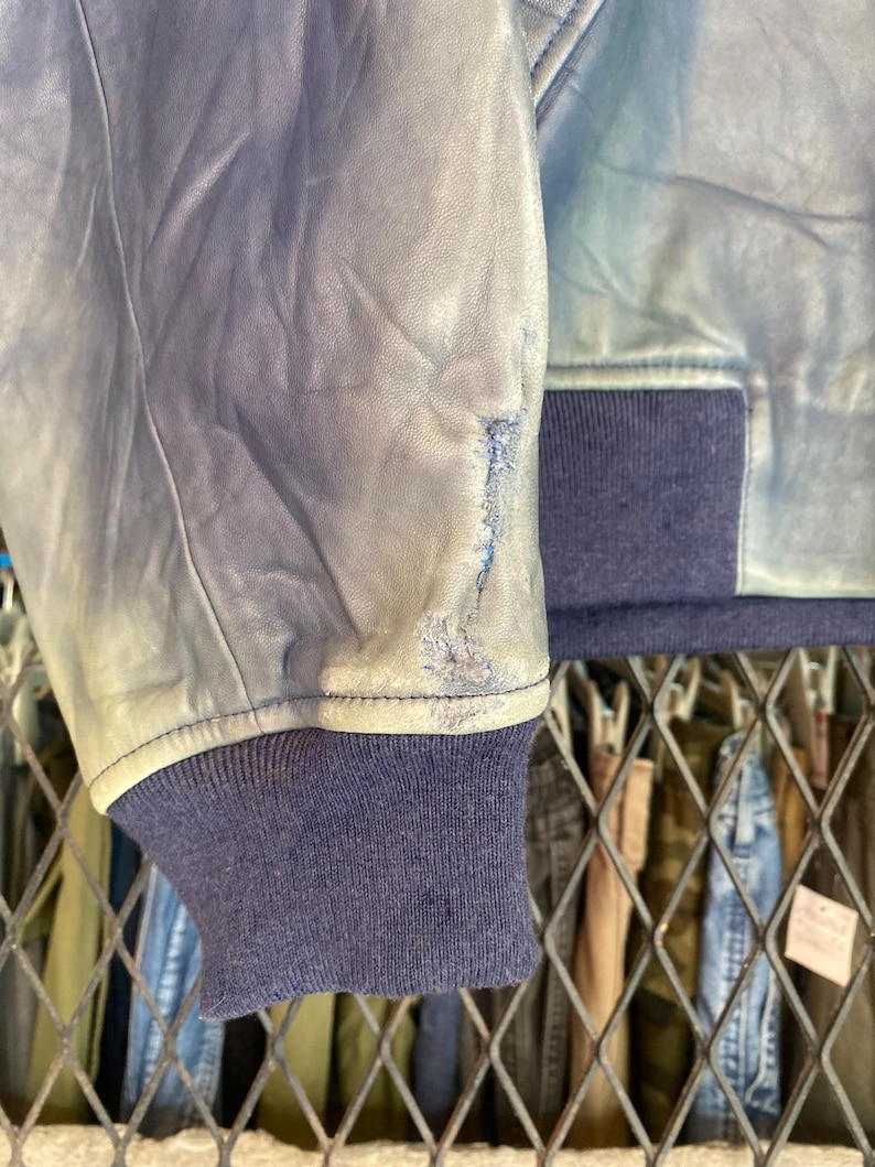 May include: Close-up of a distressed blue leather jacket sleeve. The jacket has a navy blue ribbed cuff and a light blue leather exterior with visible wear, giving a vintage look. The sleeve shows signs of wear.