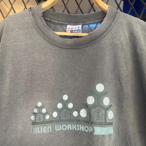 May include: A gray t-shirt with the text "ALIEN WORKSHOP" in a teal font. The shirt features a graphic of teal and white geometric shapes. The shirt is hanging on a wooden hanger.