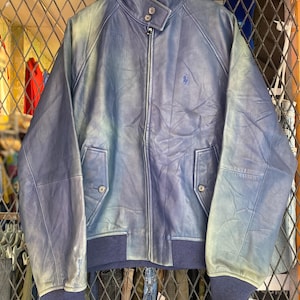 May include: A vintage blue leather jacket with a zip-up front, two front pockets, and navy blue ribbed hem and cuffs. The jacket features a small embroidered logo on the chest. The jacket is displayed on a wooden hanger.