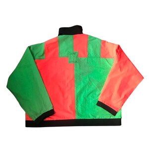 May include: A green and red color block jacket with a black zipper and a black collar. The jacket has a black band around the bottom and the sleeves. The back of the jacket has a small black square patch with the text "JAME WORLD" on it.