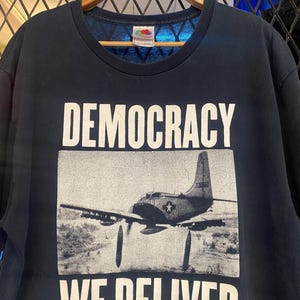 Könnte beinhalten: Schwarzes T-Shirt mit dem weißen Schriftzug "DEMOCRACY WE DELIVER" über und unter einem Graustufenbild eines Militärflugzeugs. Das T-Shirt hängt an einem Holzkleiderbügel.
