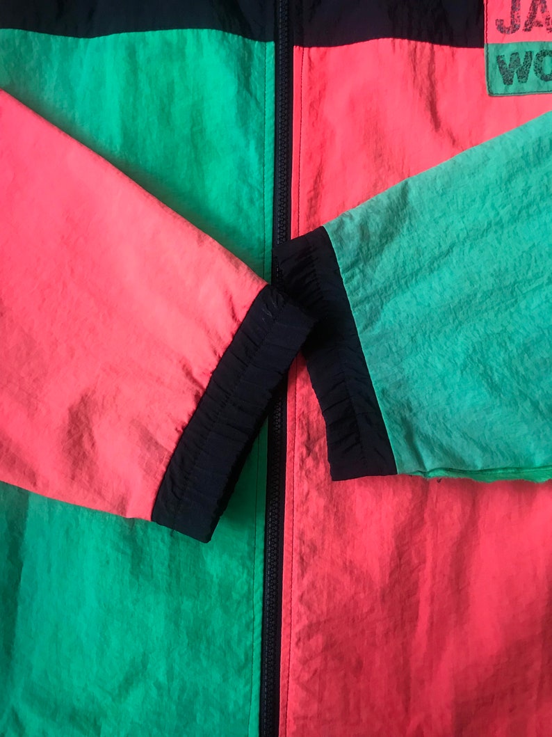 May include: A black, green, and red windbreaker jacket with a zipper closure. The jacket has a black zipper and black trim around the sleeves and collar. The text "J.A.W. WORLD" is printed on the right side of the jacket.