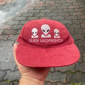 May include: Red baseball cap with the text "ALIEN WORKSHOP" and three white alien head graphics. The cap has a curved brim and is made of a textured fabric.