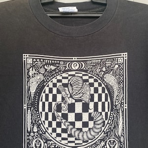 May include: Black t-shirt with a white graphic print of a butterfly and a caterpillar inside a checkered circle. The graphic is surrounded by a square frame with intricate designs and the text "MILSRNVRA" and "RC HELLCM".