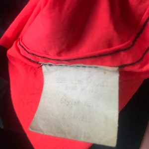May include: Close-up of a red and black jacket with a white tag that says "Soft-Line Jacket, Size: S/M, Style # 1511, Lining: 100% Polyester".