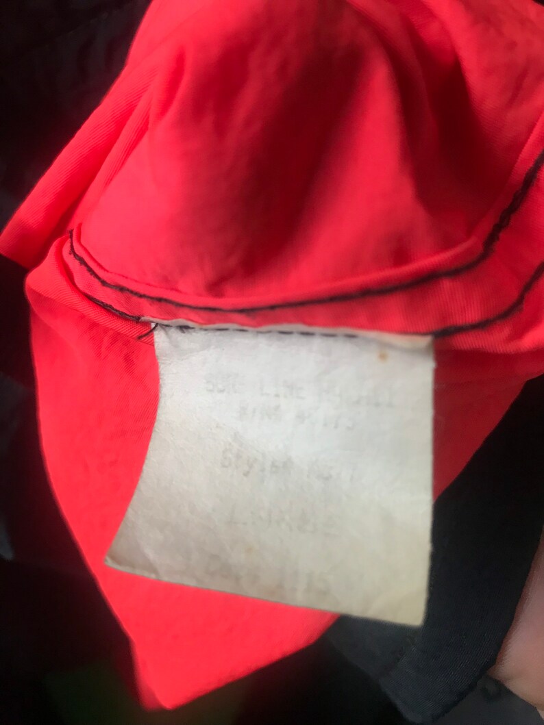 May include: Close-up of a red and black jacket with a white tag that says "Soft-Line Jacket, Size: S/M, Style # 1511, Lining: 100% Polyester".