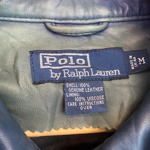 May include: A close-up of a blue leather jacket with a label that reads "Polo by Ralph Lauren." The label also indicates the jacket is made in China and is a size M. The shell is 100% genuine leather.