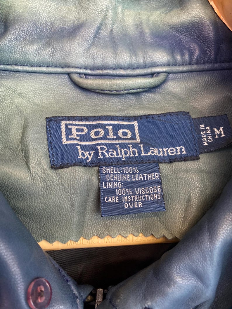 May include: A close-up of a blue leather jacket with a label that reads "Polo by Ralph Lauren." The label also indicates the jacket is made in China and is a size M. The shell is 100% genuine leather.