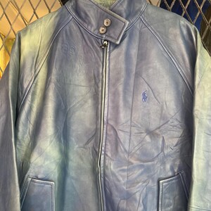 May include: A blue leather jacket with a zip-up front, two front pockets, and a stand-up collar. The jacket features a small embroidered logo on the chest and button details. The jacket is hanging on a wooden hanger.