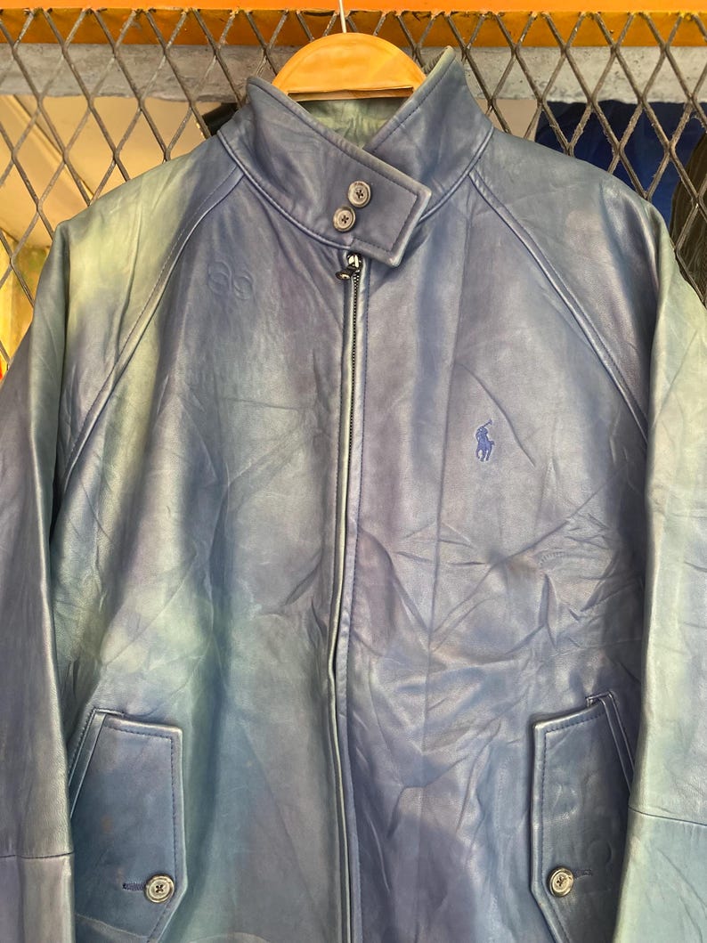 May include: A blue leather jacket with a zip-up front, two front pockets, and a stand-up collar. The jacket features a small embroidered logo on the chest and button details. The jacket is hanging on a wooden hanger.