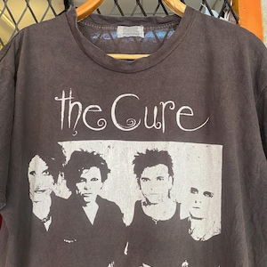 May include: A dark gray t-shirt featuring the band "The Cure" in white script above a black and white image of the band members. The shirt is hanging on a wooden hanger.