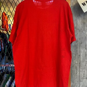 K&ouml;nnte beinhalten: Ein einfarbiges rotes T-Shirt mit Rundhalsausschnitt. Das Shirt h&auml;ngt an einem Holzb&uuml;gel.