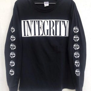 May include: Black long-sleeve shirt with the word "INTEGRITY" in large white block letters across the chest. The sleeves feature a repeating pattern of white skull graphics. The shirt is hanging on a black hanger.