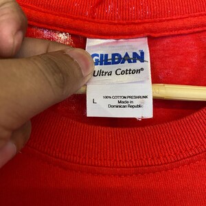K&ouml;nnte beinhalten: Rotes Gildan Ultra Cotton T-Shirt mit einem wei&szlig;en Etikett mit der Aufschrift "100% Baumwolle, vorgewaschen Hergestellt in der Dominikanischen Republik".