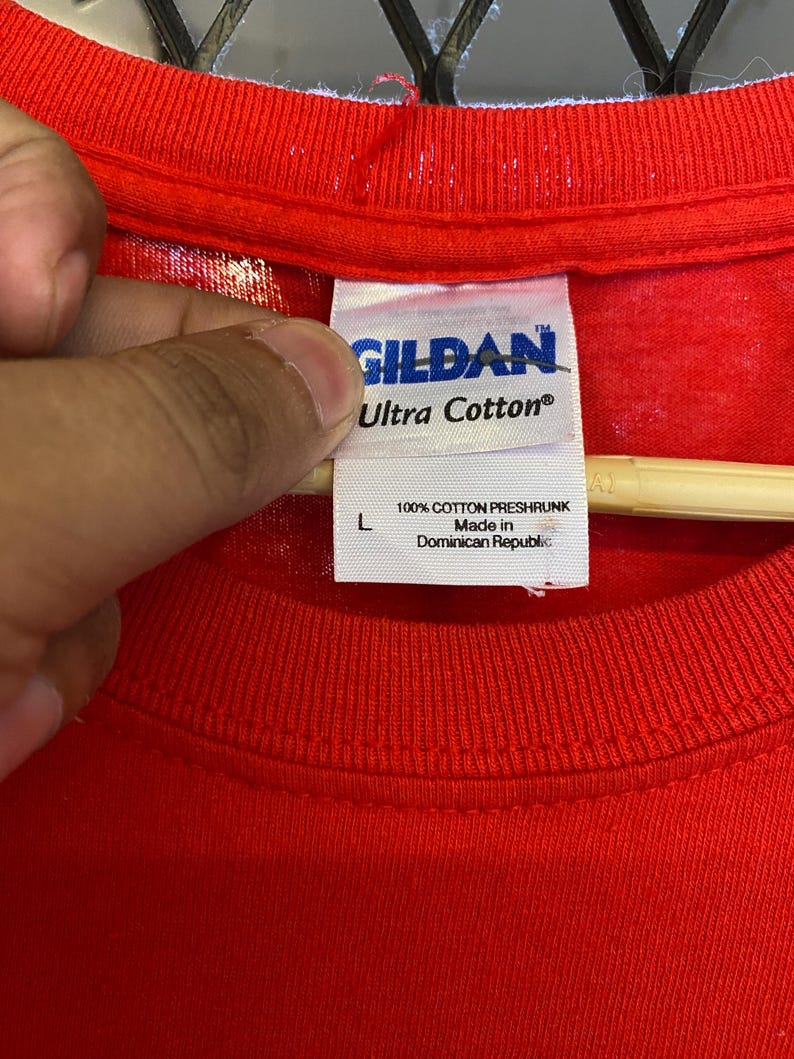 K&ouml;nnte beinhalten: Rotes Gildan Ultra Cotton T-Shirt mit einem wei&szlig;en Etikett mit der Aufschrift "100% Baumwolle, vorgewaschen Hergestellt in der Dominikanischen Republik".