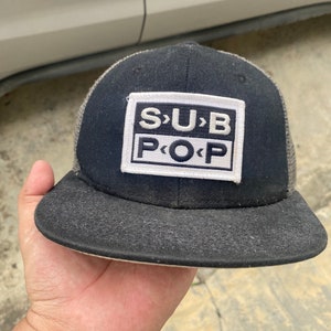 May include: A black and gray trucker hat with a white patch that says "S>U<B P<O>P".