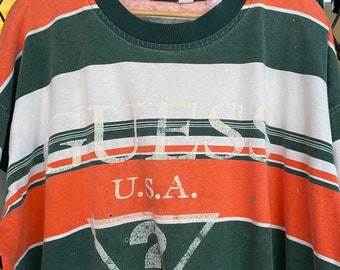 Könnte beinhalten: Ein gestreiftes Langarmshirt mit einem grünen, weißen und orangenen Farbmuster. Das Shirt trägt den Schriftzug "GUESS" in weißer Schrift und den Text "U.S.A." in weißer Schrift unter dem Wort "GUESS".