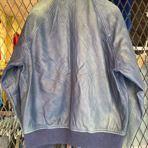 May include: A blue leather jacket featuring a stand-up collar, long sleeves, and a dark blue ribbed knit hem and cuffs. The jacket displays a smooth texture with visible natural creasing throughout the leather panels and a central back seam.