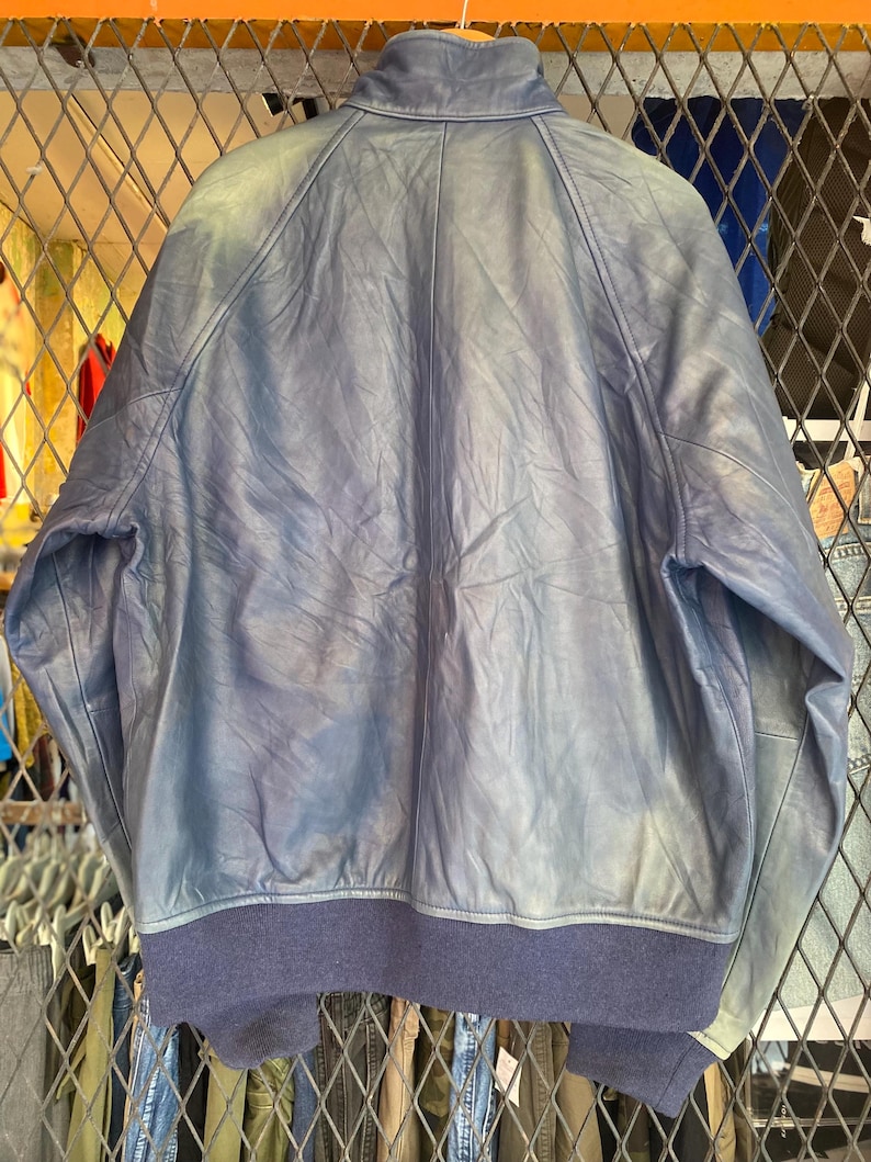 May include: A blue leather jacket featuring a stand-up collar, long sleeves, and a dark blue ribbed knit hem and cuffs. The jacket displays a smooth texture with visible natural creasing throughout the leather panels and a central back seam.
