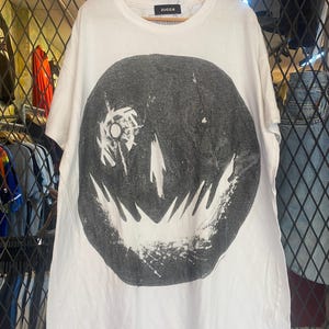 Könnte beinhalten: Weißes T-Shirt mit einem großen schwarz-weißen Grafikdruck eines Smiley-Gesichts. Der Druck ist abgenutzt und hat einen Vintage-Look. Das T-Shirt hat einen Rundhalsausschnitt und kurze Ärmel. Der Markenname "ZUCCA" ist auf dem Etikett aufgedruckt.