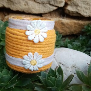 Peut inclure: Un pot décoratif enveloppé de corde jaune, orné d'appliqués de marguerites blanches et d'un ruban rose clair. Le pot est posé sur un fond de succulentes vertes et d'un mur en pierre, créant une esthétique rustique et artisanale.