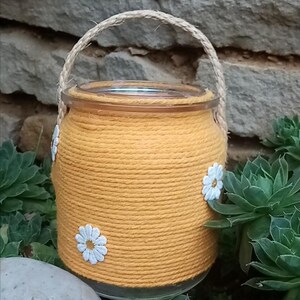 Peut inclure: Un bocal en verre jaune enveloppé de ficelle, orné de marguerites blanches. Le bocal a un couvercle en verre transparent et une poignée en corde tressée. Le fond présente de la verdure et un mur de pierre.