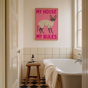 K&ouml;nnte beinhalten: Ein rosa-wei&szlig;es Poster mit einer Katzenillustration und dem Text "My House My Rules". Das Poster h&auml;ngt an einer wei&szlig;en gekachelten Wand in einem Badezimmer mit einer wei&szlig;en Badewanne und einem Holzhocker.