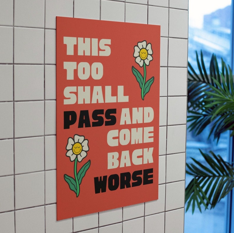 Pu&ograve; includere: Un poster rosso con testo bianco che recita "This Too Shall Pass And Come Back Worse". Il poster presenta due fiori bianchi con facce sorridenti gialle.