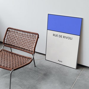 May include: A brown wicker chair with a metal frame sits in front of a white and blue poster with the text "Rue de Rivoli, Paris".