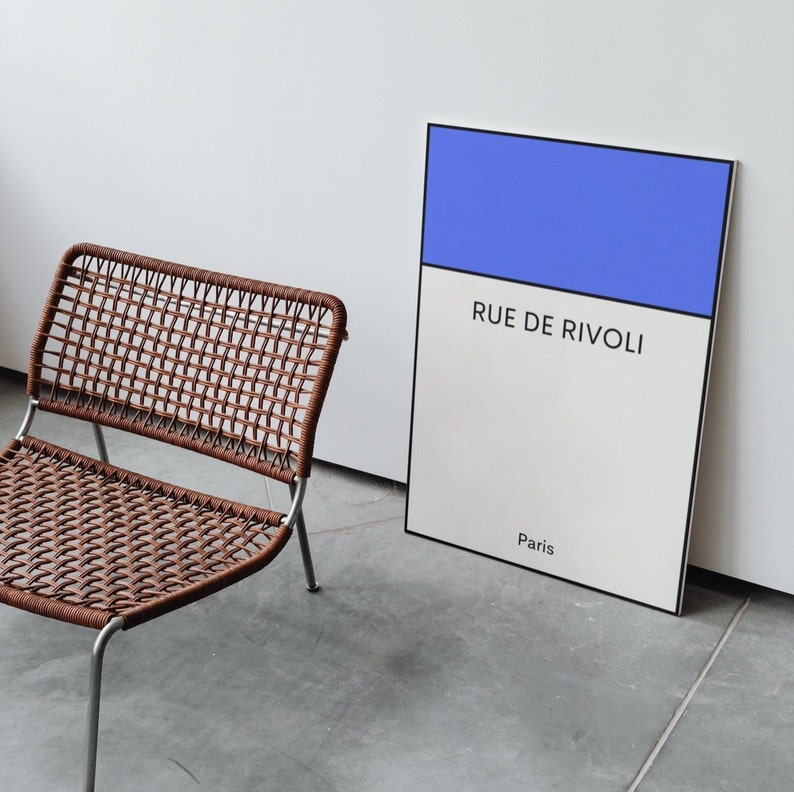 May include: A brown wicker chair with a metal frame sits in front of a white and blue poster with the text "Rue de Rivoli, Paris".