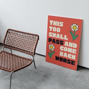 Pu&ograve; includere: Un poster rosso con il testo "This Too Shall Pass And Come Back Worse" in lettere bianche e nere. Il poster presenta due fiori bianchi con centri gialli e facce sorridenti.