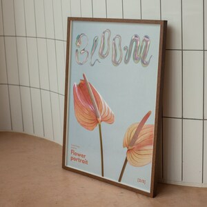May include: A framed print with a light blue background featuring the word "bloom" in iridescent letters. Two pink and orange anthurium flowers are depicted in the print. The text "Flower portrait" is printed below the flowers. The print is labeled "[2/5]".
