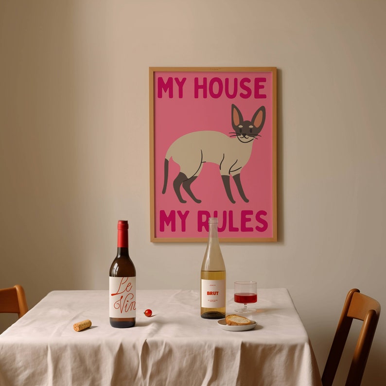 K&ouml;nnte beinhalten: Ein rosa gerahmtes Poster mit einer wei&szlig;en Katze und dem Text "My House My Rules". Eine Flasche Wein, ein Glas Wein und ein Korken befinden sich auf einer wei&szlig;en Tischdecke.