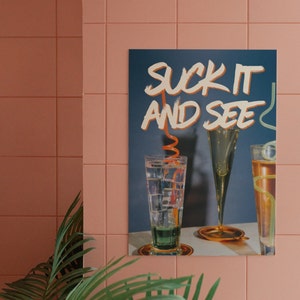 May include: A blue poster with white text that reads "Suck It And See". The poster features three glasses with orange straws, two of which are filled with a clear liquid and one with a dark liquid.