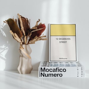 May include: A beige ceramic vase with a wavy design holds a bouquet of dried brown and red flowers. The vase is sitting on a white surface in front of a stack of books with the text "Mocacfico Numero" and "12 Degraves Street, Melbourne".
