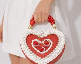 May include: A heart-shaped red handbag with white frosting-like trim and pearl accents. The bag features three cherry embellishments and a red handle with gold-colored hardware.