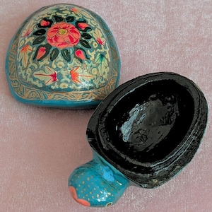 Könnte beinhalten: Kleine, ovale Schmuckschatulle mit Klappdeckel. Der Deckel ist mit einem floralen Muster in Rosa, Grün und Gold auf türkisfarbenem Grund bemalt. Der Sockel ist türkis mit goldenen Akzenten, das Innere ist schwarz.