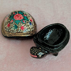 Könnte beinhalten: Dekorative, schildkrötenförmige Schmuckschatulle. Der Deckel zeigt ein Blumenmuster mit roten, rosa und türkisfarbenen Akzenten auf cremefarbenem Hintergrund. Die schwarze Basis hat einen gepunkteten Kopf und einen rosa Mund.
