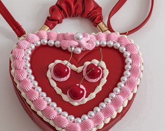 May include: A red heart-shaped handbag with a white and pink frosting design and three red cherries. The bag has a red strap and a red handle.