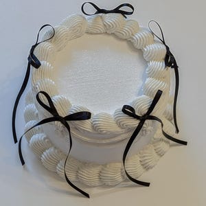 Op de afbeelding: Een witte cake met zwarte lintjes rond de randen. De cake is versierd met witte frosting en heeft een glinsterend wit oppervlak.