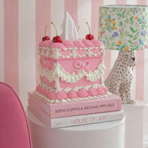 Könnte beinhalten: Eine rosa Taschentuchbox in Kuchenform, verziert mit Kirschen und Zuckergussdetails. Die Box steht auf zwei Büchern, mit den Titeln "SOFIA COPPOLA ARCHIVE 1999-2023" und "HOUSE OF JOY". Eine dekorative Lampe und eine Dalmatinerstatue befinden sich im Hintergrund.