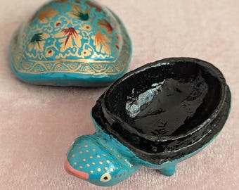 May include: A turquoise turtle-shaped trinket box with a black interior and a matching lid. The lid is decorated with gold floral patterns and red accents. The turtle's head has a pink mouth and gold polka dots.