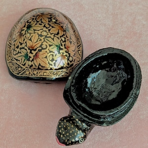 Könnte beinhalten: Dekorative, offene schildkrötenförmige Schmuckschatulle. Der Deckel ist schwarz mit einem goldenen Blumendesign. Der Körper der Schildkröte ist schwarz mit goldenen Akzenten und einem rosa Mund. Das Innere der Box ist schwarz.