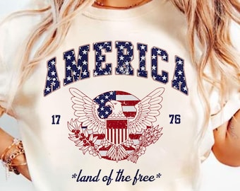 May include: A white t-shirt with a vintage-style graphic of an eagle with outstretched wings holding a shield with the American flag. The text "AMERICA" is above the eagle and "17 76" is on either side of the eagle. The text "*land of the free*" is below the eagle.