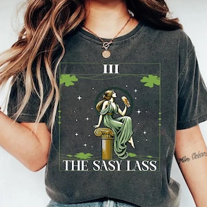 May include: A dark gray t-shirt with a graphic of a woman sitting on a pillar, holding a glass of beer. The graphic is surrounded by shamrocks and stars. The text "III THE SASy LASS" is printed below the graphic.